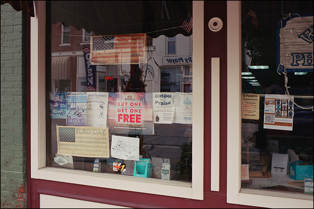 Signs in the front window of Warren Pharmacy in the small town of Warren, Indiana. There are two paper American flags, a couple of advertisements for products sold in the store, and several signs for religious events and churches.