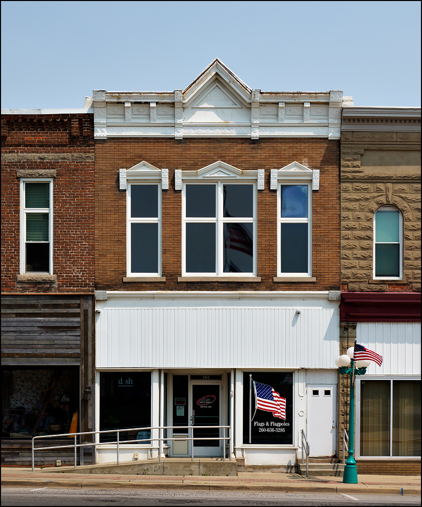 Ebey Sales Flags and flagpoles storefront in a beautiful old brick commercial building on Orange Street in the small town of Albion, Indiana. An American flag flies from a light pole in front of the building and another large American flag is reflected in the second floor windows.