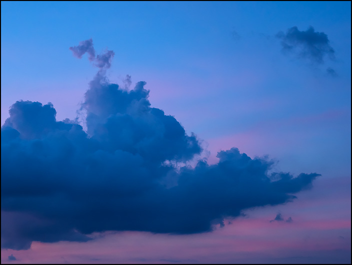 An abstract photograph of a red and purple sky at dusk with a big blue cloud that is shaped like a spearhead.