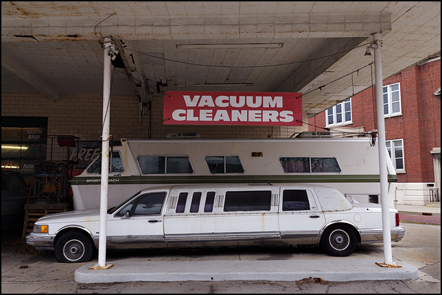 A beat-up old Lincoln Town Car stretch limousine with flat tires and an old motorhome are parked under a banner advertising vacuum cleaners in front of Allen County Sweeper Company on Broadway in Fort Wayne, Indiana.