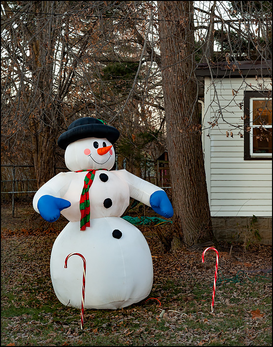 An inflatable snowman and large plastic candy canes stand in the front yard of a house on Arbor Avenue in Fort Wayne, Indiana. Fall leaves cover the ground around them.