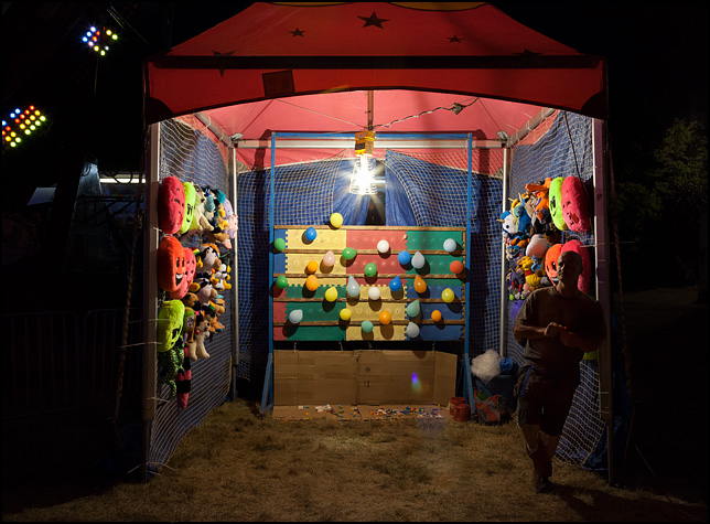 A carny leans against the tent pole next to the balloon-dart game he runs at the Three Rivers Festival carnival in Fort Wayne, Indiana. The tent has stuff animals hanging on the sides and a board with balloons on the back wall.