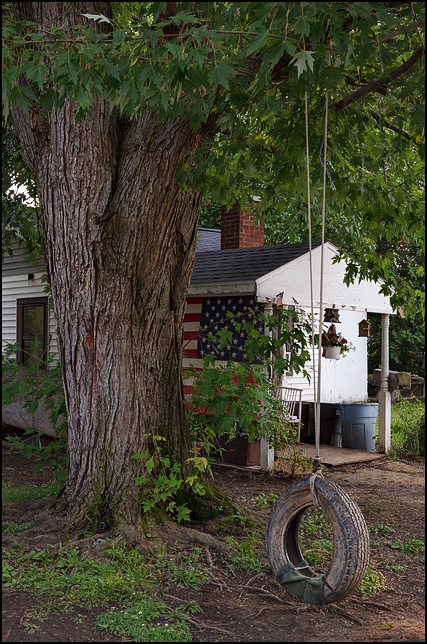An old tire swing hanging from a tree in front of a small house on Huron Street in a working class neighborhood in Fort Wayne, Indiana. A large American flag covers the side of the front porch.