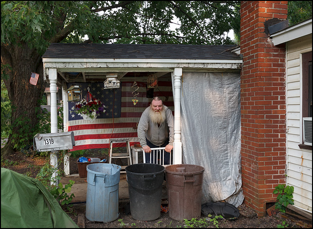 An old man with a big beard stands in front of a large American flag on the front porch of his house on the working-class west side of Fort Wayne, Indiana.