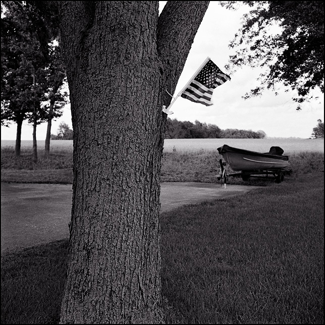 A little American flag hangs on the side of a tree on State Road 18 in rural Blackford County, Indiana. A boat on a trailer is visible in the background in front of a field.