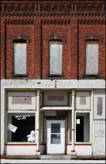 A vacant storefront in a two story brick building on Broadway in the small town of Butler, Indiana. A plush toy snowman sits in the empty window. A sign saying that the store has moved is painted on the glass.