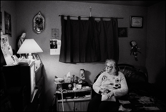An elderly woman sits in her bedroom opening the mail and watching television. She is a devout Catholic with crosses and statues of the Virgin Mary all over the walls. She also has two Betty Boop dolls.