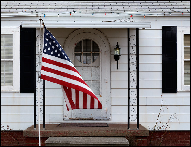 An American flag hangs limp in front of a house as the wind begins to blow.