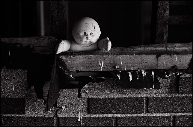 A Cabbage Patch Kid doll looking out through the broken wall of an abandoned house.