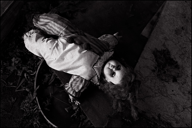 A clown doll with closed eyes laying on the floor of an abandoned house surrounded by trash.