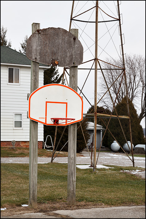 Two basketball backboards, one above the other, next to a farmhouse on State Road 37, near the state line, in rural northeast Allen County, Indiana. One of the backboards is an old one of weathered wood, and the hoop is missing. The other is a modern one with a hoop.