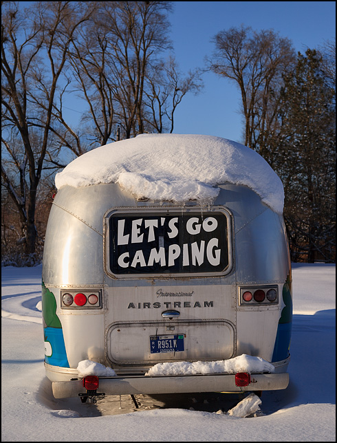 An old Airstream travel trailer with a decal over the rear window that says Lets Go Camping. The camper sits in deep snow during the winter in Indiana.