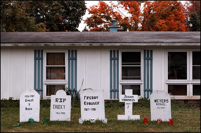 A house decorated for Halloween with plywood tombstones in the yard with the names of horror movie killers Michael Myers, Jason Voorhees, and Freddie Krueger. Skeletons are crawling out of the ground in front of the gravestones.
