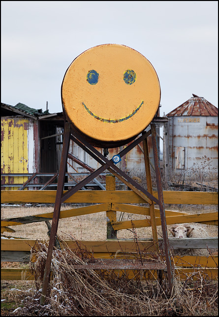 A fuel tank with a yellow happy face painted on the front of it stands in front of a barnyard fence in rural Delaware County, Indiana. A sheep looks out through the fence.