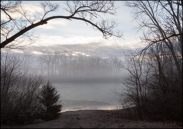 Fog at sunrise hangs over the pond at Hodell Acres, the WMCA summer camp on Elmhurst Drive in Fort Wayne, Indiana. The surface of the pond is covered in a thin layer of ice. A view through the trees that surround the deep depression where the pond, a former quarry, is located.
