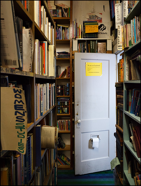 A Facebook sign hangs on the inside of the restroom door, above the toilet paper roll, at Hyde Brothers Books in Fort Wayne, Indiana. Seen looking down an aisle of old books.