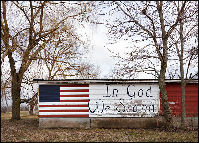 An American flag with no stars painted on a barn next to a sign that says In God We Stand. The barn is on South County Line Road in rural Allen County, Indiana.