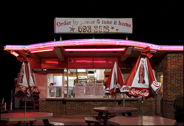 The carry-out window at Magic Wand Restaurant in Churubusco, Indiana. Photographed at night with the neon lights turned on. Tables with Coca-Cola umbrellas provide seating in front of the restaurant.