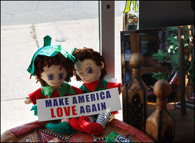 Two dolls holding a bumper sticker that says, Make America Love Again. They are sitting on a table in Little Shop of Lauras, a secondhand shop on Broadway in Fort Wayne, Indiana.