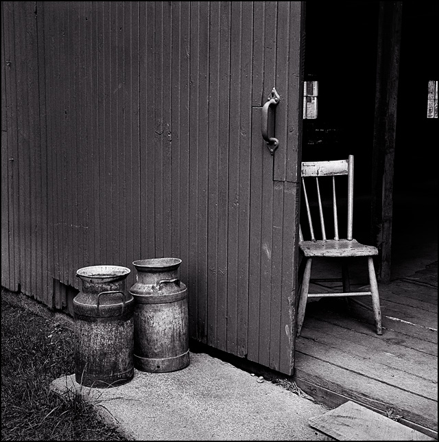 An old wooden chair sits inside a barn, and two rusty antique milk cans sit outside next to the door.