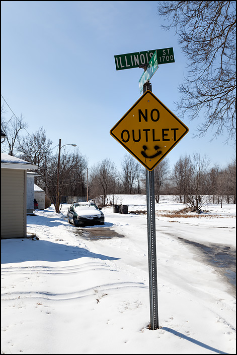 A yellow road sign that says No Outlet, located on the corner of Manufacturers Avenue and Illinois Street in the West Main Street neighborhood in Fort Wayne, Indiana. There is a graffiti drawing of a smiley face at the bottom of the sign.
