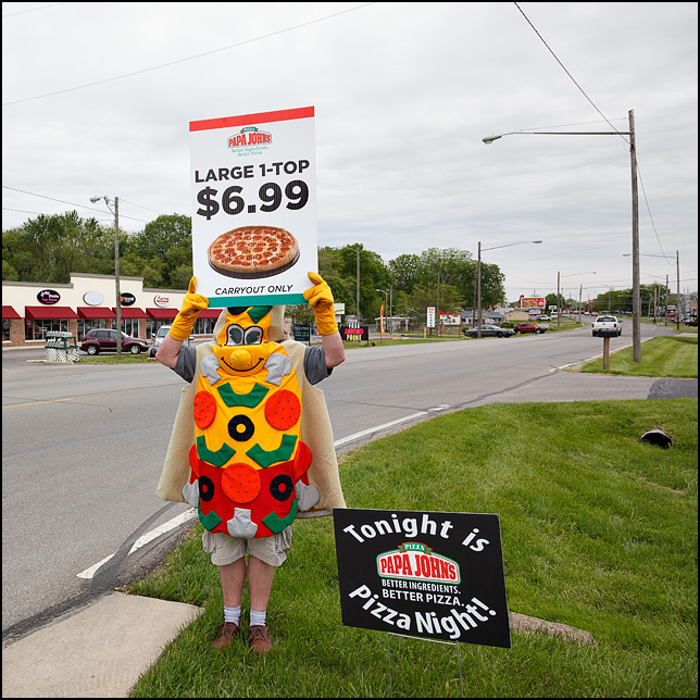 A man wearing a Pizza Slice costume while holding a Papa Johns sign on Bluffton Road in the Waynedale area of Fort Wayne, Indiana.