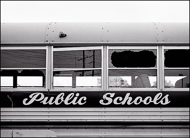 An old school bus with broken windows that has Public Schools painted on the side instead of the name of the school district.