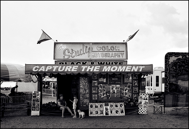 A little girl and her dog chat with the woman who runs the photo booth at the Rodeo de Santa Fe carnival. The booth has a big sign that says "Capture the moment", and the outside is covered in framed portraits.