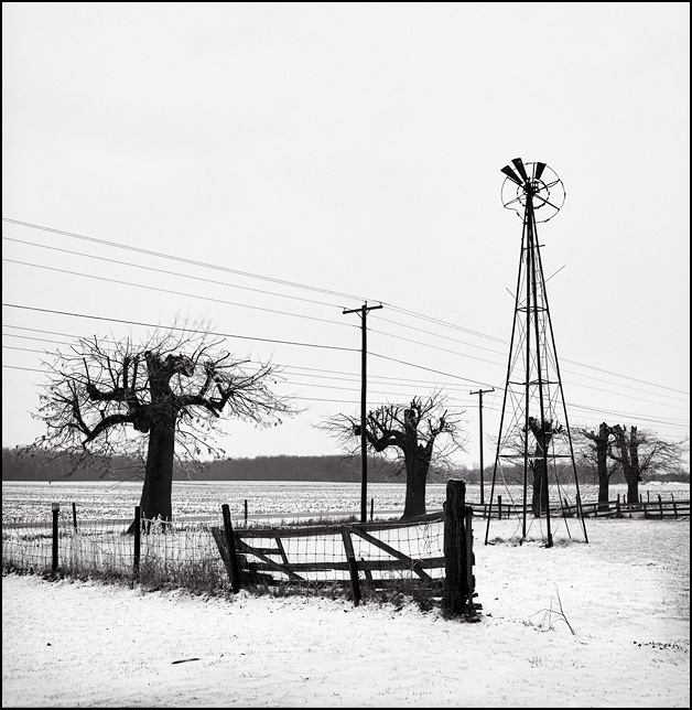 An old rusty windmill in an abandoned barnyard with a broken fence, all covered in snow during the cold Indiana winter.