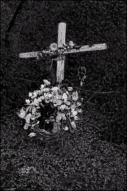 A large wooden roadside memorial cross adorned with a floral wreath stands in the woods along Smith Road in Fort Wayne, Indiana.