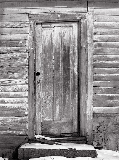 Snow covers the ground under the weathered wood back door of an abandoned farmhouse on Till Road in rural northwest Allen County, Indiana. The door and siding are covered in peeling white paint.
