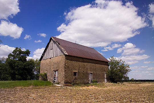 Old abandoned cinderblock barn with wood and metal roof in rural Pulaski County, Indiana. The deep blue sky is filled with dramatic fluffy white clouds. The barn sits in an overgrown lawn next to an empty cornfield.