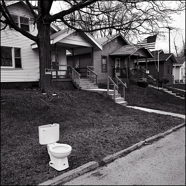 A toilet left out for the garbage collection sits on the curb in front of a house in Fort Wayne, Indiana. An American flag flies in front of another house in the distance.