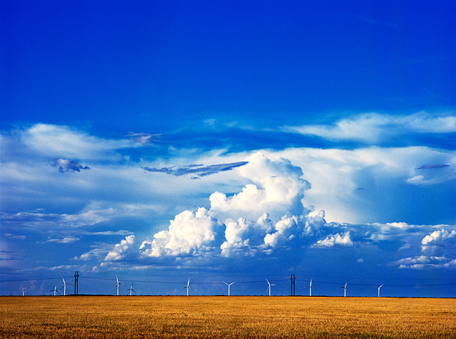 Windmills stand under a big blue sky and huge white clouds on a farm in Vega, Texas.