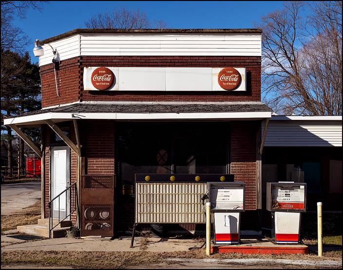 An abandoned gas station in the small town of Tyner, Indiana. Two old gas pumps stand in front of the brick building, and there are antique Coca-Cola signs above the windows. 