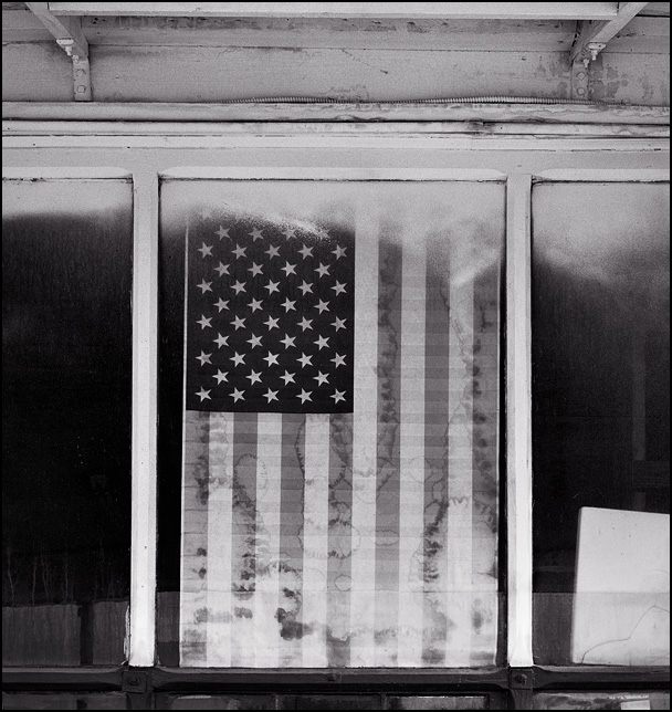 A faded paper American flag covered in mildew hangs in the front window of a small town auto repair shop in Uniondale, Indiana.
