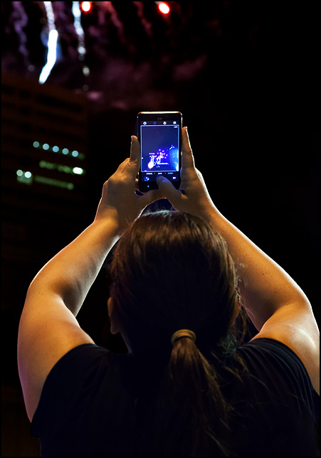 A woman holding up her smartphone at the 2017 Fourth of July Fireworks in Fort Wayne, Indiana. She is watching the fireworks on the screen.