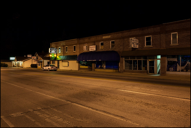 A night photograph of the main business district of Waynedale on Lower Huntington Road in Fort Wayne, Indiana. The Elzey building with the Waynedale Bakery, Hook and Ladder Tavern, and Meyers Barbershop are visible along the road.