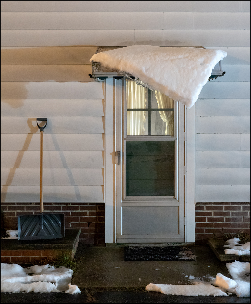 A sheet of snow on the metal awning over the back door of my house has partially slid off, hanging off the front of the awning like a blanket hanging off the side of a bed. A snow shovel stands next to the door.