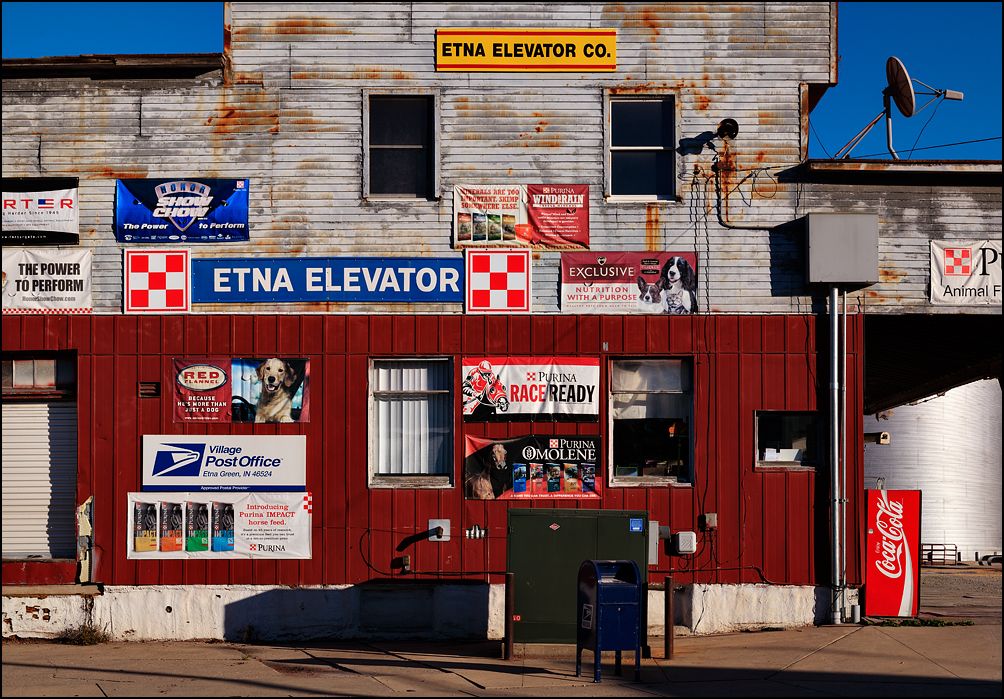The front of the Etna Elevator animal feed store in the small town of Etna Green, Indiana. The weathered wood building has several signs for feed companies like Purina, and a US Postal Service sign.