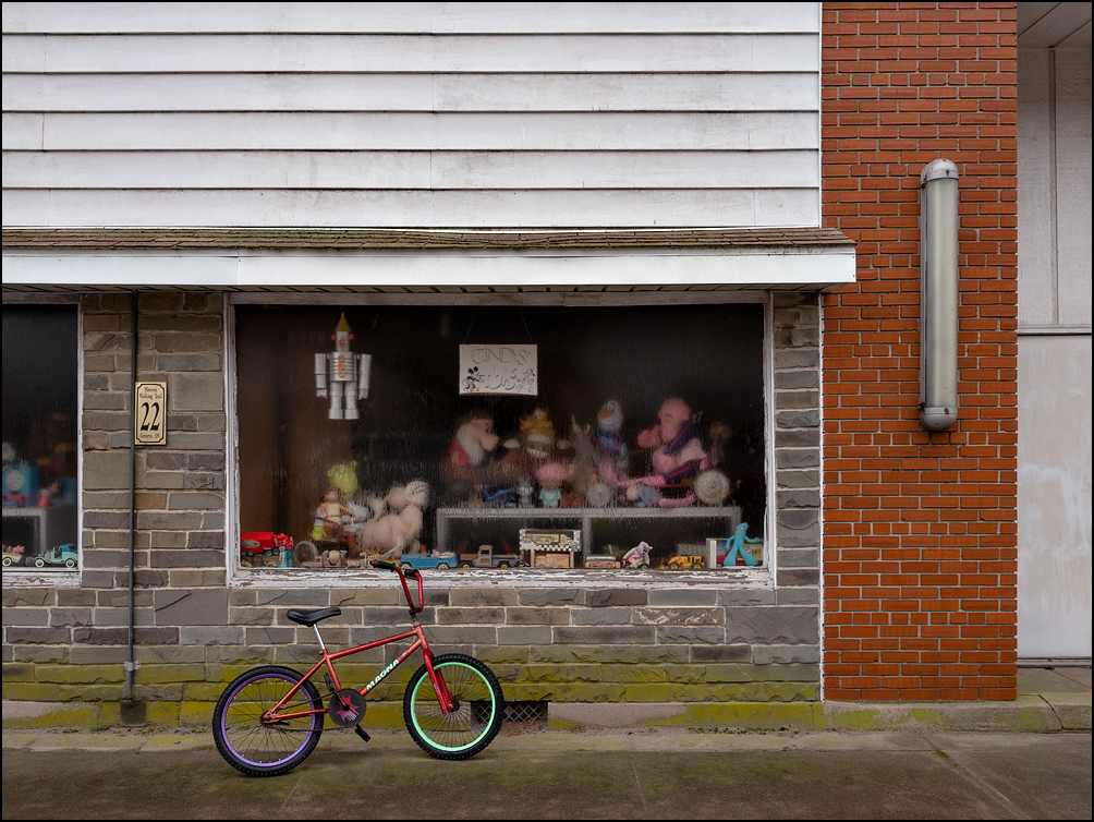 A childs BMX bicycle leaned up against the front of a building on a rainy day in the small town of Geneva, Indiana. The large windows on the storefront on Line Street are filled with old toys and a handwritten sign in the window says Cindys Toys.