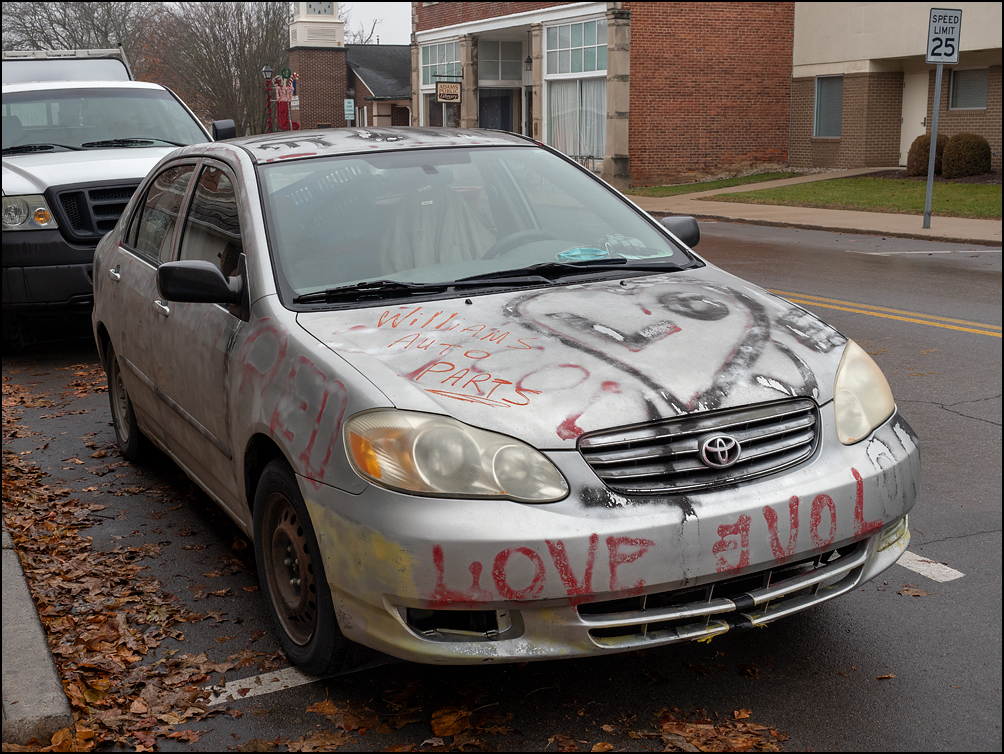 An old beat up Toyota car covered in spray painted graffiti of hearts and love parked on Line Street in the small town of Geneva, Indiana.