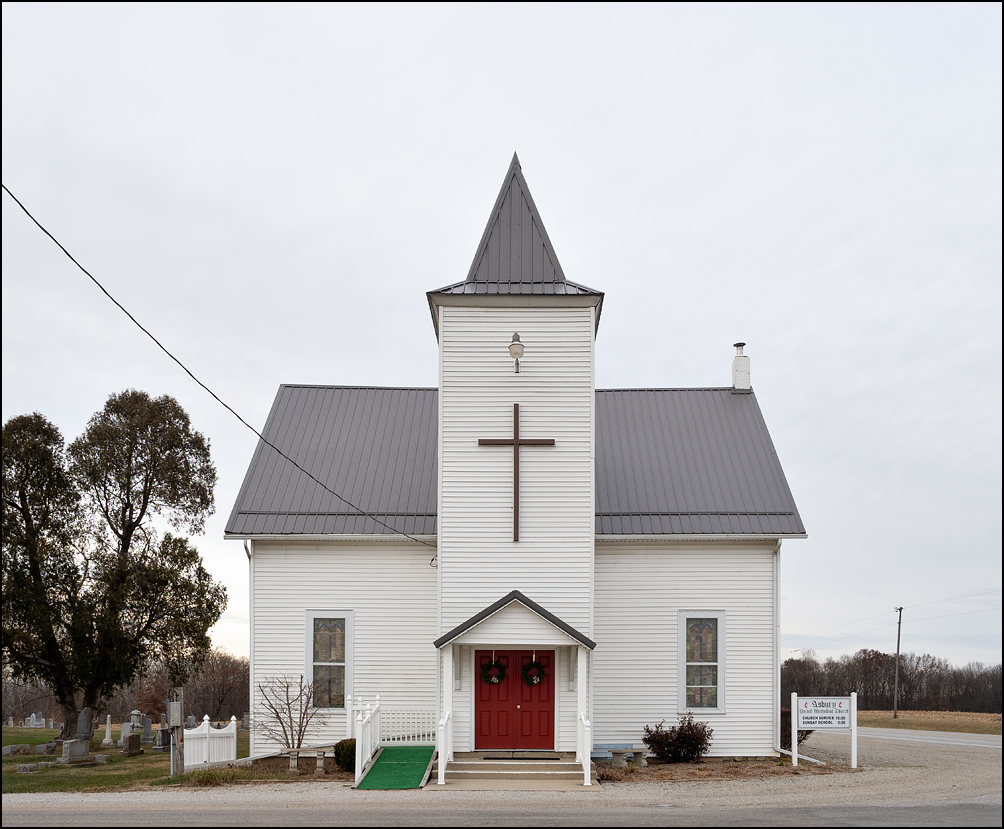 Asbury United Methodist Church, a small white church surrounded by a cemetery on County Road 1100S in rural Wells County, Indiana. A large cross hangs on the front of the church above the red doors, which are decorated with Christmas wreaths.