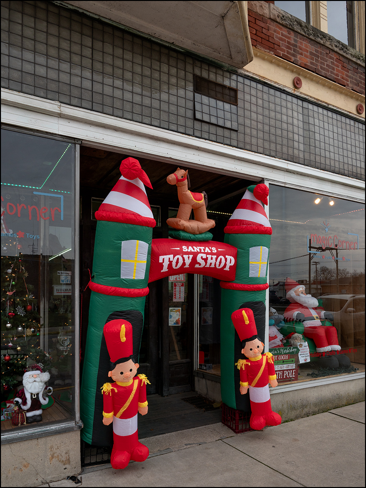 An inflatable arch that says Santas Toy Shop at the entrance of a store called Magic Corner on Washington Street in the small town of Hartford City, Indiana. The display windows have an inflatable Santa Claus and Christmas trees.