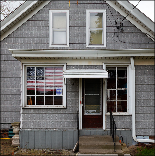 A large American flag covers a window on the back of an old house, and a sticker on the door warns that burglars will be killed.