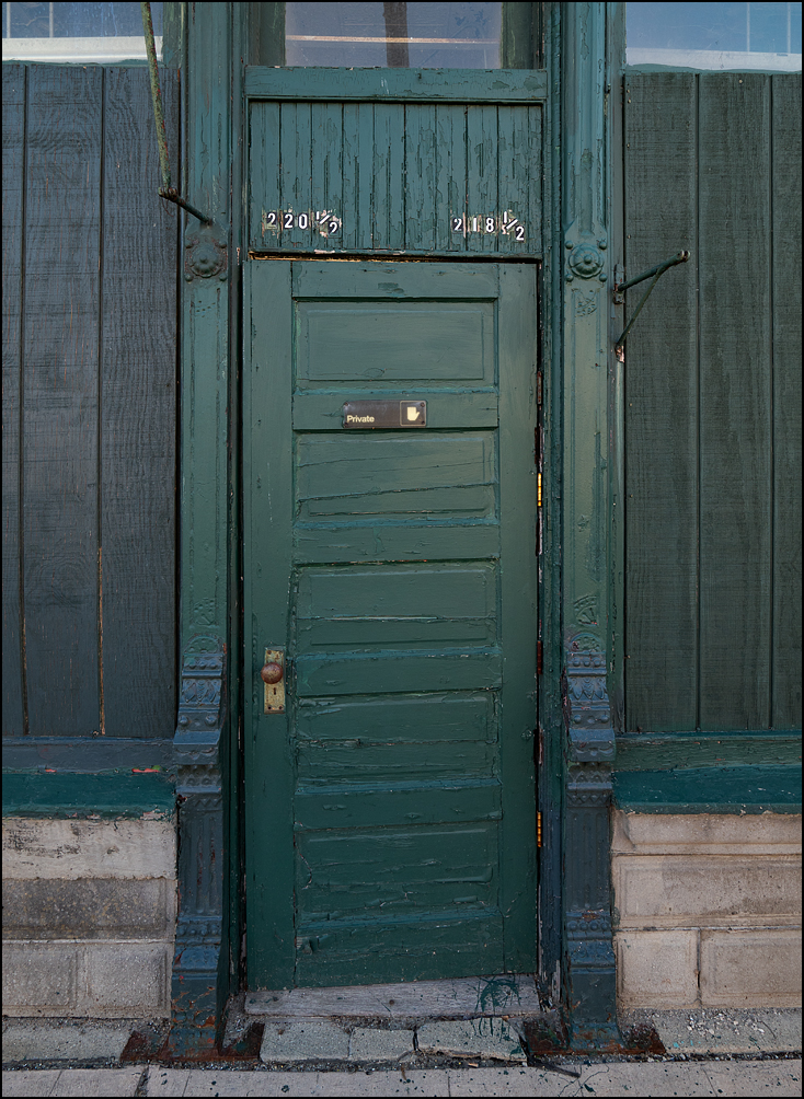 An old green panel door with an ornate cast-iron frame in a boarded-up storefront on Third Street in the small town of Burr Oak, Michigan. A sign on the door says Private.