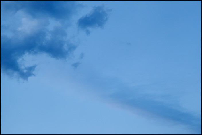 An abstract photograph of a long cloud pointing at a cluster of dark clouds in a blue sky on a June evening in the small town of Churubusco, Indiana.