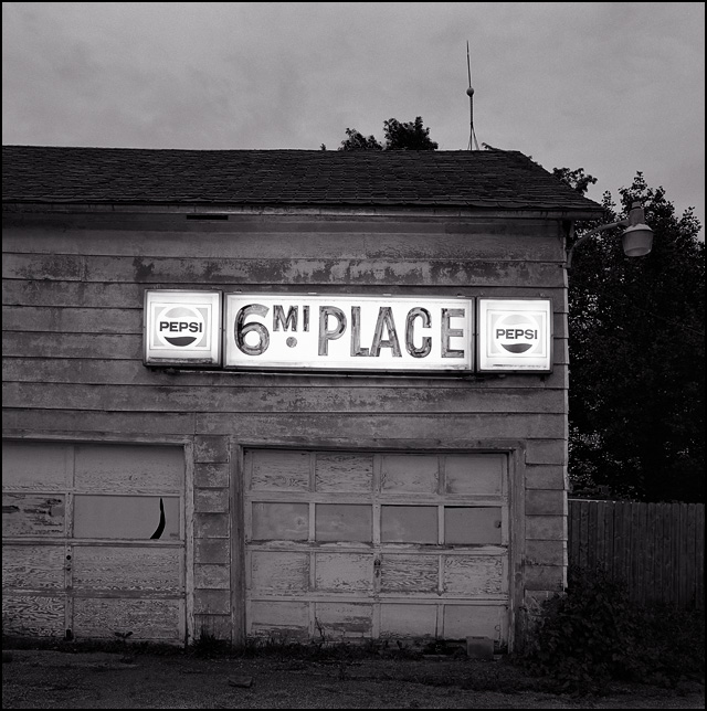 The lit-up sign for 6 Mile Place Pizzeria and Bar on Wayne Trace outside Fort Wayne, Indiana. The sign has Pepsi logos on it and it hangs on a rundown old garage behind the bar.