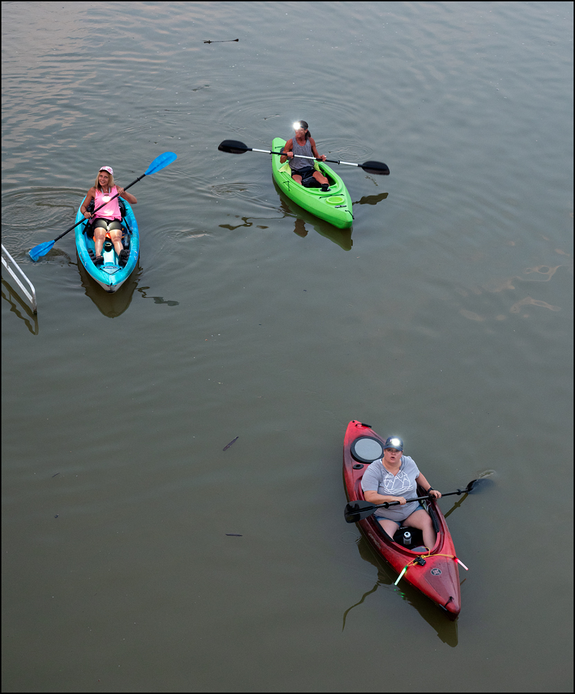 Three women in colorful plastic kayaks on the Saint Marys River at Promenade Park in downtown Fort Wayne, Indiana. Photographed in the evening near dusk and the women are wearing headlamps.