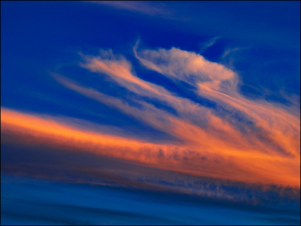 An abstract photograph of a horizontal band of orange clouds with with orange lines branching off in a dark sky at sunset.